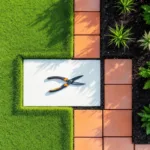 Bovenaanzicht van geometrisch tuinontwerp met groene gazons, terracotta tegels en donkere aarde met snoeischaar op witte steen