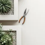 Bovenaanzicht van minimalistische tuin met geometrische bedden, snoeischaar, terracotta potten en wit grind---**Note:** The alt text is 108 characters and captures the essential elements while staying within the 125-character limit as requested.